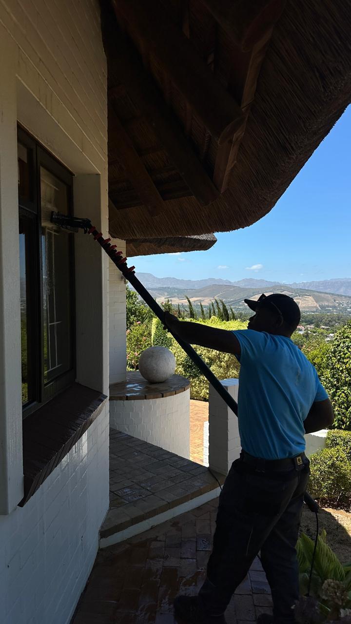 Blu cleaner washing windows under a thatched roof with vineyards and mountains behind