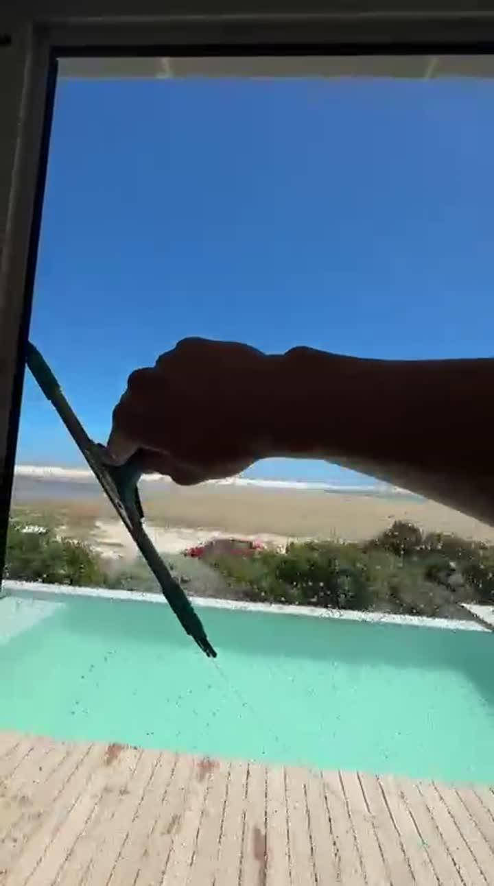 Squeegee cleaning a glass pane beside a pool, with the beach behind